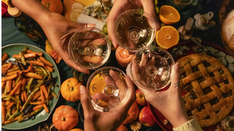 op view of a festive Thanksgiving table with roasted vegetables, mini pumpkins, pie, and four hands raising glasses in a toast, celebrating togetherness and gratitude.